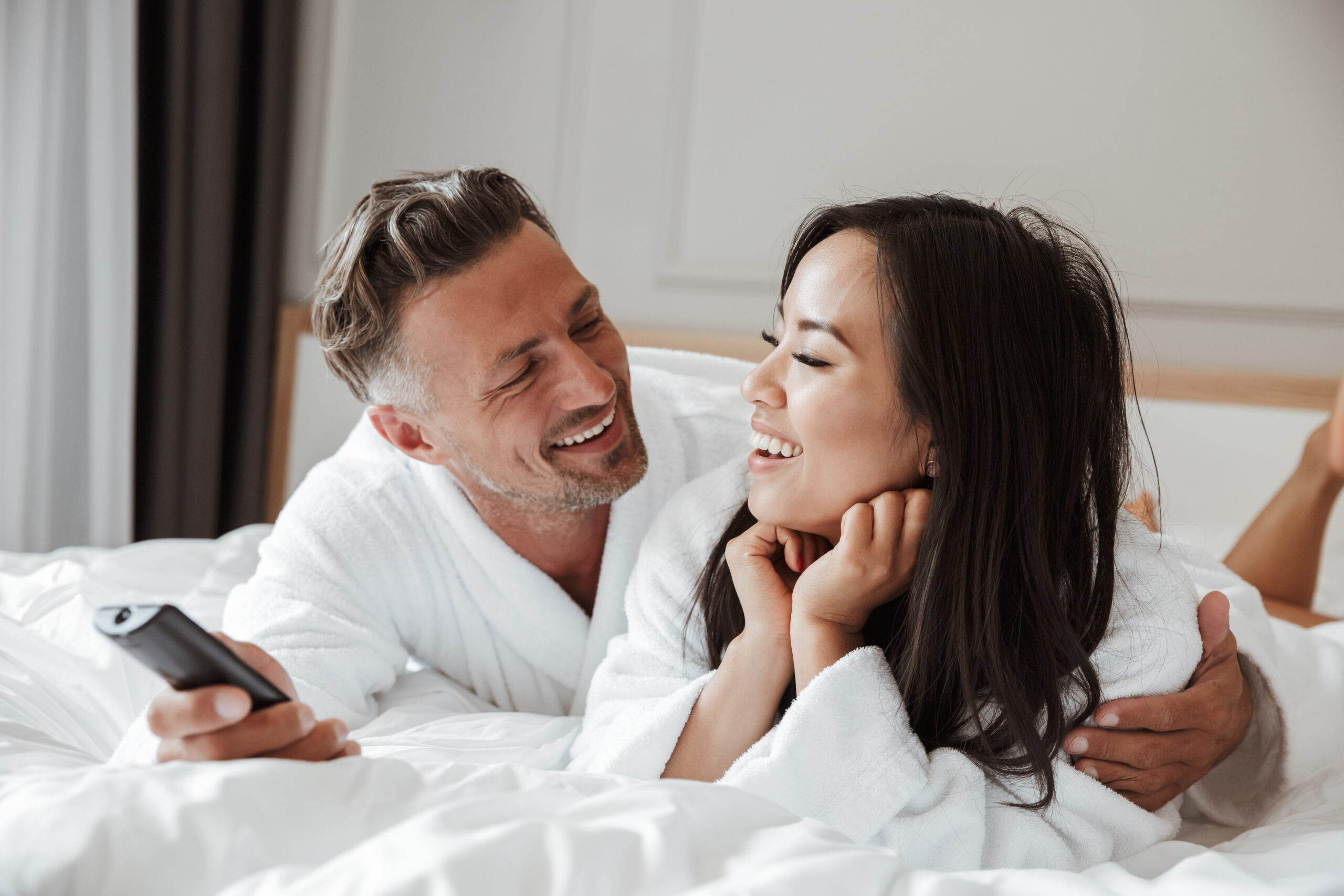 Multiethnic couple caucasian man and asian woman wearing white housecoat lying in home bedroom apartment with remote control in hand while changing tv channels